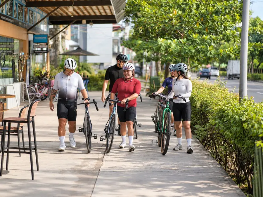 Four cyclists walking their bikes along a tree-lined path in the morning